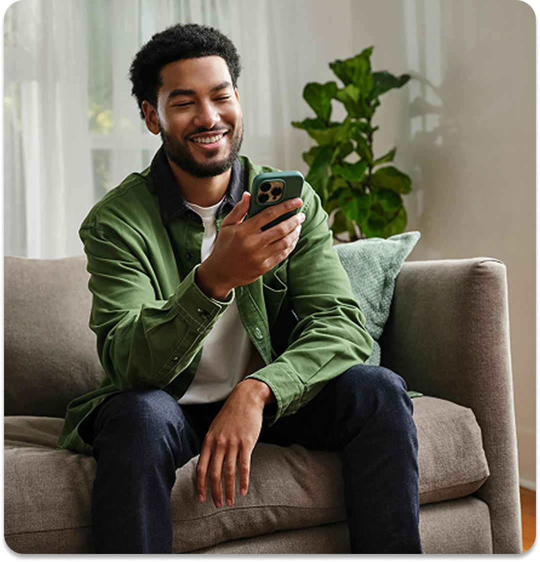 An adult male sitting on a couch smiling at his phone.
