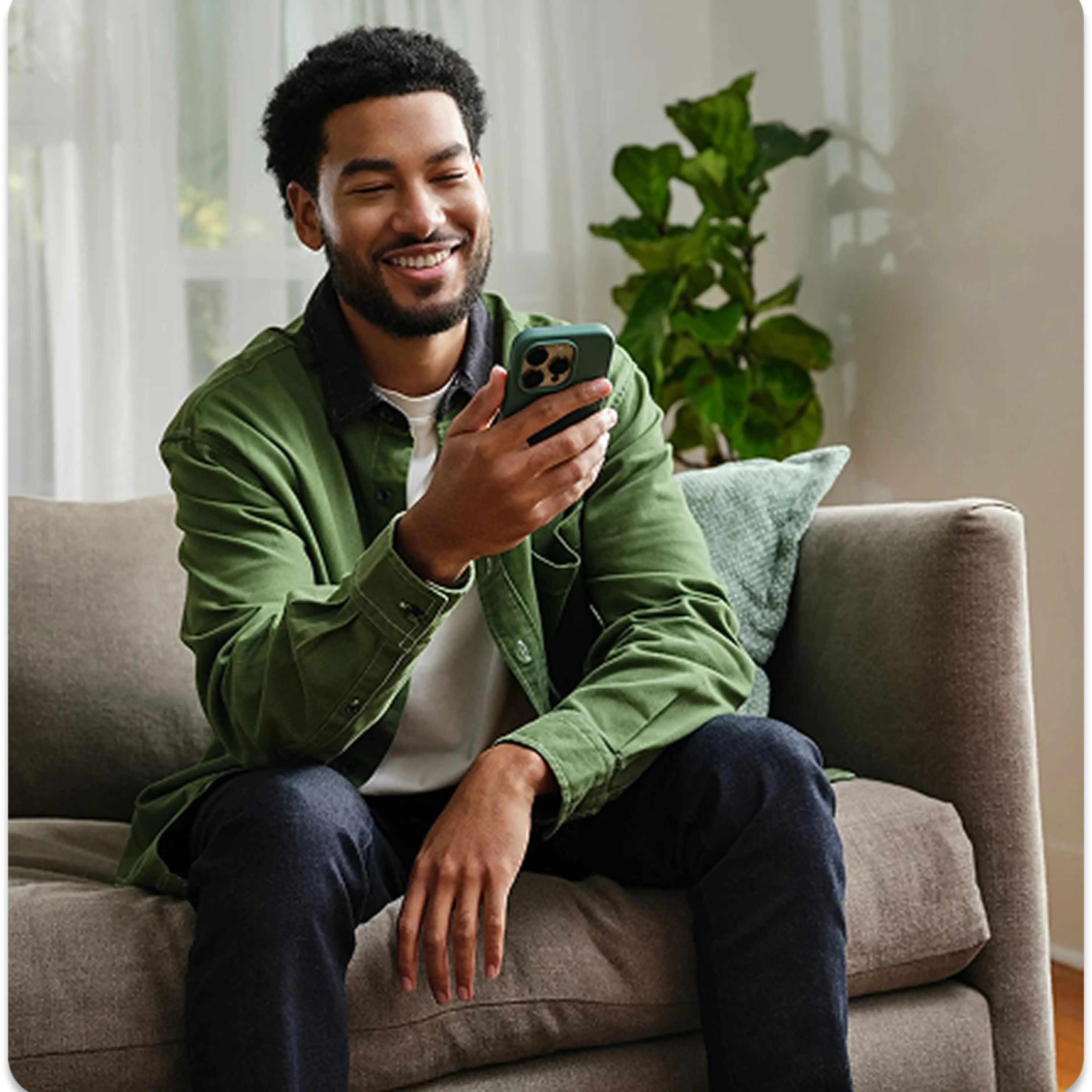 An adult male sitting on a couch smiling at his phone.